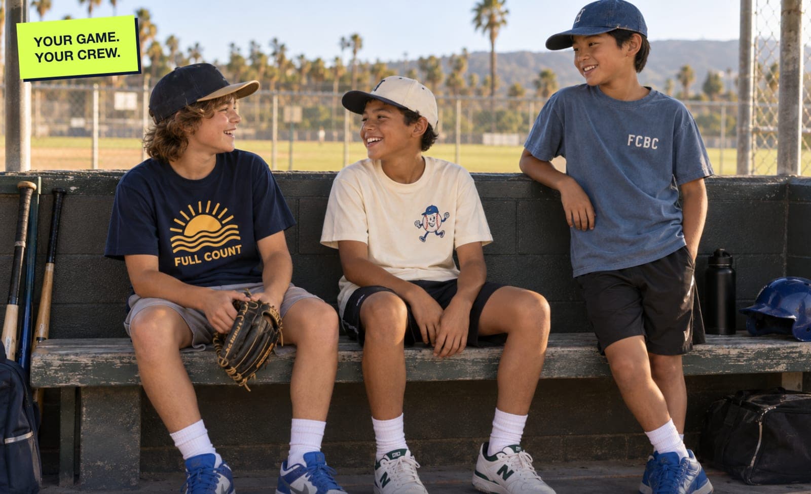 Three kids in FCBC tees laughing on a dugout bench under the palms